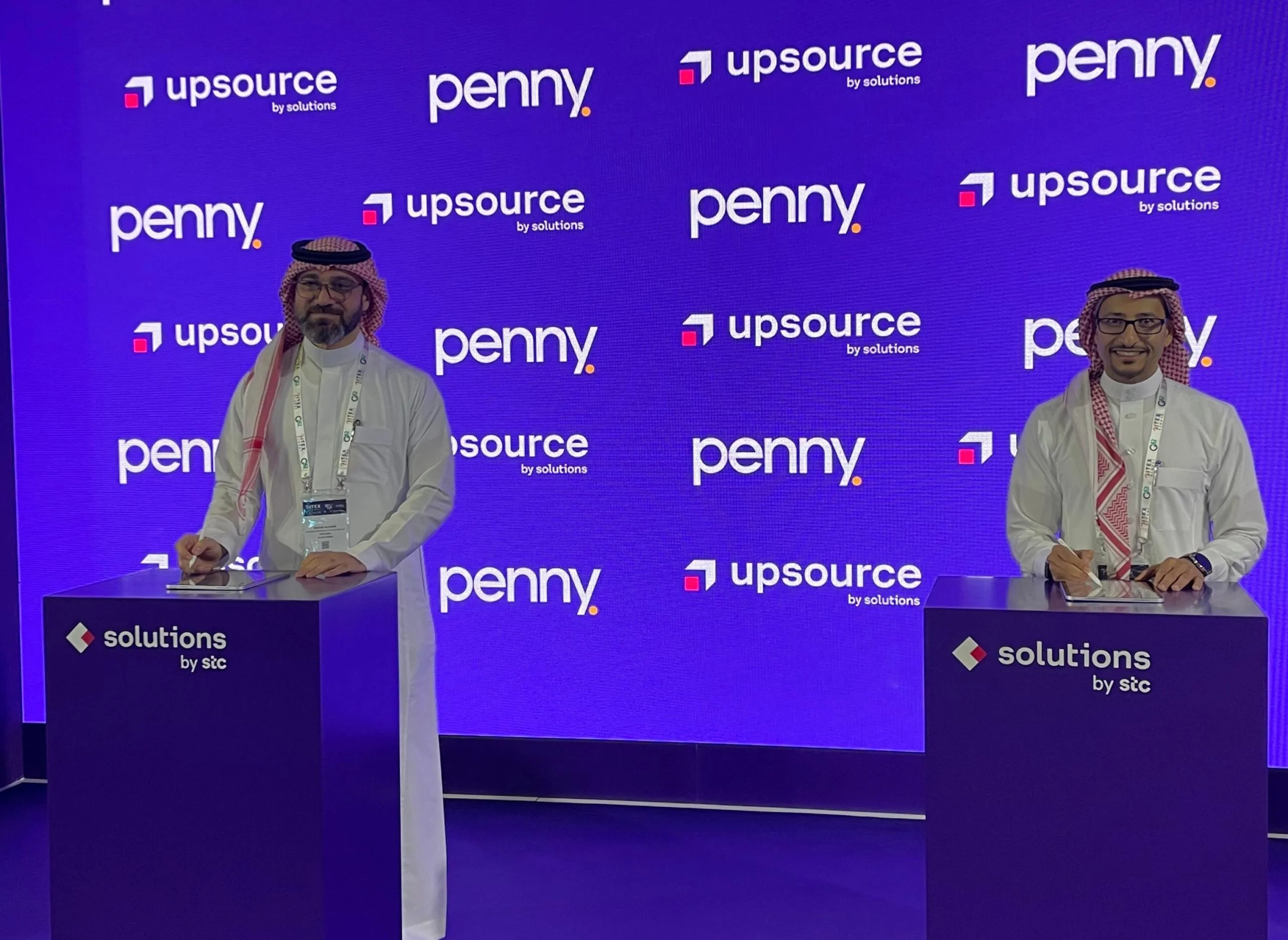Two business executives in traditional Saudi attire at corporate signing ceremony with Penny and Upsource branding